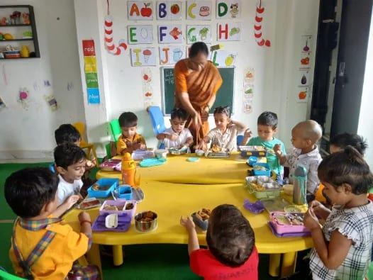 Children eating from tiffin boxes around a low table while a teacher serves
