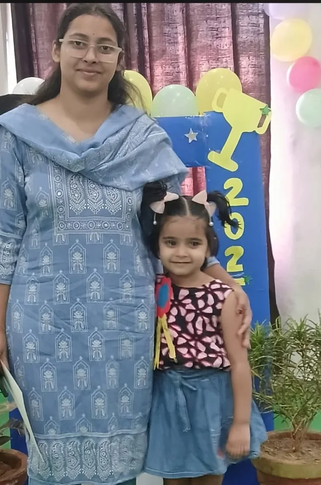 A teacher standing with a young child beside a decorated backdrop and potted plants