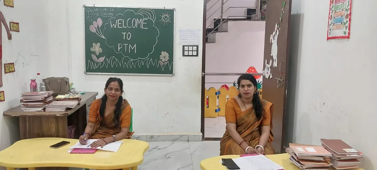 Two teachers seated at desks with registers and papers beneath a hand-drawn 'Welcome to PTM' board