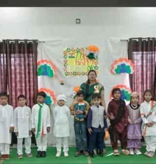 Children in white and tricolour dress standing on a decorated school stage in front of a hand-made banner
