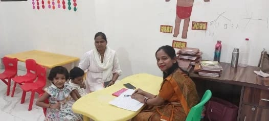 A teacher writing at a desk with registers and notebooks stacked beside her while two children sit at a low table