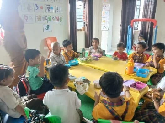 Children seated around a table with tiffin boxes, alphabet and number charts on the wall behind