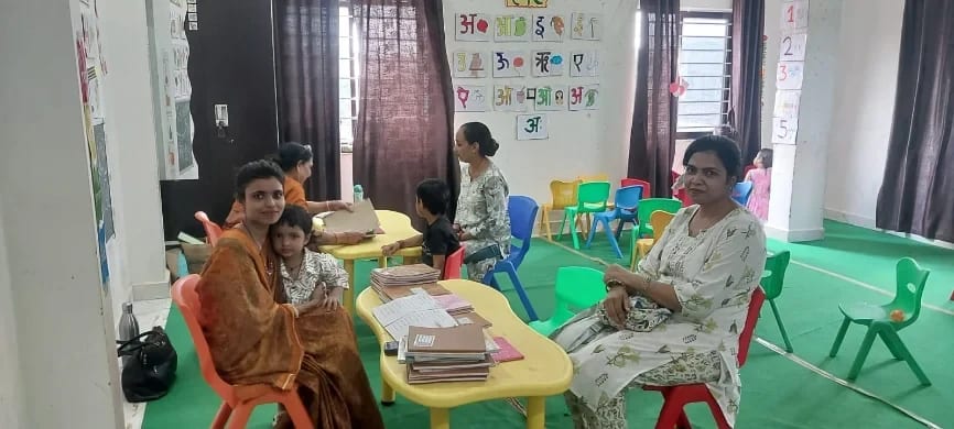 A classroom with Hindi alphabet and number charts on the wall, low tables stacked with notebooks, and teachers seated with parents and children