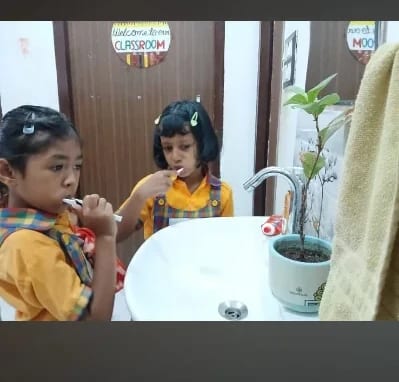 Two children brushing their teeth at a washbasin beneath a hand-made 'Welcome to our classroom' sign