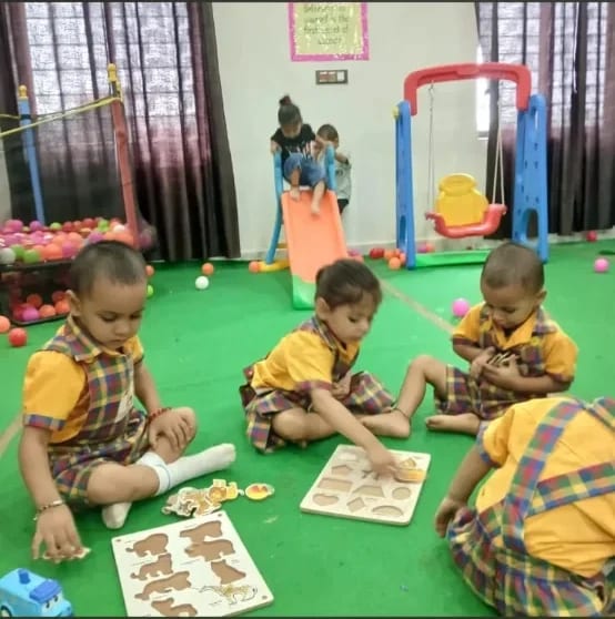 Children seated on the floor of the activity room fitting shapes into wooden puzzle boards, with a slide and ball pit behind them